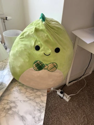 Stor grön Squishmallow med fluga - Supermjuk och stor grön Squishmallow i form av en drake  med vit mage och söt grönrutig fluga. Perfekt som kramdjur eller mysig inredningsdetalj. Tillverkad i mjuk plysch och har ett gulligt ansikte med broderade detaljer.
