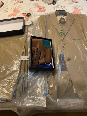 Kostymset från Riley - Hej! Jag säljer ett helt nytt kostymset från märket Riley. Setet är aldrig använt eller ens provat då det var en present i fel storlek.  Passform: Slim fit  Storlek:  * Kavaj: 48 * Byxor: 48  Färg: Travertine (ljus/beige ton)  Ingår i setet:  * Slips * Näsduk * Strumpor * Manschettknappar Fluga  Allt är helt nytt.  Nypris:  * Kavaj: 1800 kr * Byxor: 800 kr * Tillbehör: 500 kr  Totalt nypris: ca 3100 kr Mitt pris: 1500