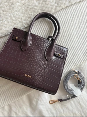 Burgundy croco handväska från ALDO - Snygg mörklila handväska från ALDO i croco-mönstrat konstläder. Väskan har två handtag, guldfärgade detaljer och medföljande axelrem. En matchande liten rund väska med kedja ingår också. Perfekt accessoar för att lyfta din outfit. Nypris 800kr.