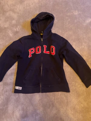 Marinblå hoodie från Polo Ralph Lauren - Snygg marinblå hoodie från Polo Ralph Lauren med dragkedja och stor röd POLO-text på bröstet. Klassisk huva och två fickor framtill. Tillverkad i mjuk bomullsblandning som känns skön mot huden. Perfekt för en avslappnad och sportig stil.