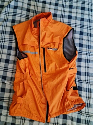 Orange funktionsväst från Peak - Orange funktionsväst från Peak med svarta meshpaneler på sidorna, reflexdetaljer och flera fickor. Västen har hög krage, dragkedja framtill och justerbara band. Perfekt för utomhusaktiviteter när du vill synas.