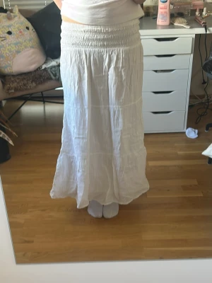 Vit långkjol i linne - Säljer en vit långkjol med smock i midjan och lätt, flowigt tyg. Kjolen har en avslappnad passform och är perfekt för varma dagar. Tillverkad i linne för en luftig känsla och stilren look.