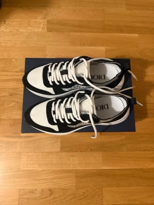 Dior B22 vita och svarta sneakers - Snygga Dior B22 sneakers i vitt och svart med klassiskt Dior-mönster på sidan. Skorna har en chunky vit sula, svarta och vita paneler i mesh och mocka samt vita skosnören. Perfekta för dig som vill sticka ut med exklusiva detaljer och modern design.