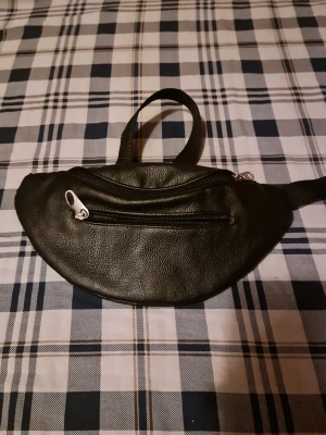 Svart magväska i skinnimitation - Snygg svart magväska i skinnimitation med två dragkedjefack. Perfekt att bära runt midjan eller över axeln. Enkel och stilren design som passar till många outfits.