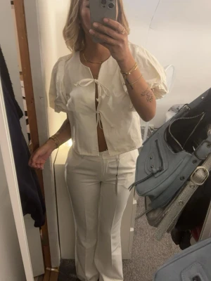 Vit blus med knyt och puffärm - Trendig vit blus med kort ärm och puffiga axlar. Blusen har två knyt framtill och är croppad, vilket ger en modern och avslappnad look. Perfekt att styla med höga byxor eller kjol. Materialet känns lätt och luftigt.