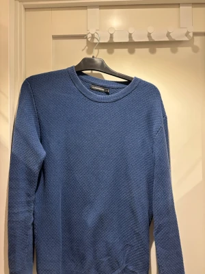 Blå  tröja från J.Lindeberg | strl M - Snygg blå stickad tröja från J.Lindeberg med rund halsringning och diskret strukturmönster. Perfekt för lager-på-lager och enkel att matcha med jeans eller chinos. Tröjan har långa ärmar och ribbade muddar.