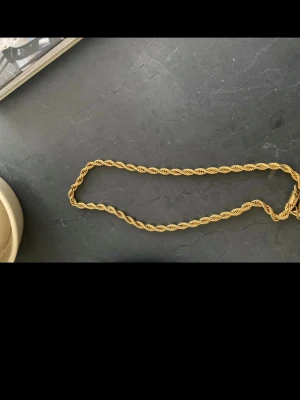 Guldfärgat tvinnat halsband - Säljer ett stilrent guldfärgat halsband med tvinnad kedja. Halsbandet har en klassisk design och är tillverkat i metall med en snygg glansig finish. Perfekt för dig som gillar minimalistiska accessoarer med lite edge.