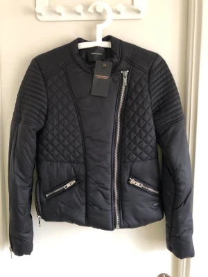 Svart jacka från Maison Scotch - Snygg svart pufferjacka från Maison Scotch med quiltade detaljer på axlar och sidor. Jackan har asymmetrisk dragkedja, två snedställda fickor med dragkedja och ribbade partier vid ärmslut och midja. 