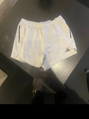 Grå träningsshorts från Adidas med prislapp kvar - Snygga ljusgrå träningsshorts från Adidas med elastisk midja och svarta detaljer vid fickorna. Perfekta för sport eller gym och tillverkade i ett lätt och bekvämt polyestermaterial. Adidas-logga på ena benet ger en sportig look.