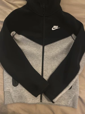 Nike tech grå svart (helt ny) - Nike hoodie med dragkedja i svart och ljusgrått. Klassisk design med vit Nike-logga på bröstet, huva och två fickor framtill. Tillverkad i mjukt material som är skönt att ha på sig. Perfekt för en sportig och avslappnad stil. 