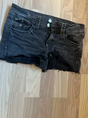 Svarta jeansshorts från Gina Tricot - Snygga svarta jeansshorts från Gina Tricot med råa kanter och slitna detaljer. Shortsen har klassisk femficksdesign, dubbla knappar i midjan och vita kontrastsömmar. Perfekta för en avslappnad stil under varma dagar.