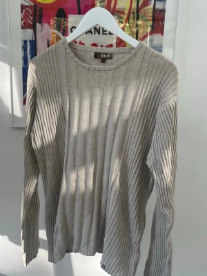 Beige ribbstickad tröja från Emilio - Snygg beige ribbstickad tröja från Emilio med rund halsringning och långa ärmar. Tröjan har en klassisk och stilren look med vertikala ränder som ger en modern känsla. Perfekt att matcha med jeans eller kjol för en avslappnad stil. Har ett hål på höger armbåge enligt bild. 