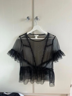 Svart meshblus med volanger H&M - Säljer en transparent svart meshblus från H&M med volangdetaljer på ärmar och midja. Blusen har rund halsringning och korta ärmar med rysch. Perfekt att styla över ett linne eller bralette för en trendig look.