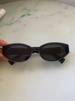 Svarta cat eye-solglasögon från Miu Miu - Snygga svarta solglasögon från Miu Miu i trendig cat eye-modell. Glasögonen har mörka linser och breda skalmar med guldiga Miu Miu-loggor på sidorna. Ramen är i blank plast som ger en cool och modern vibe.
