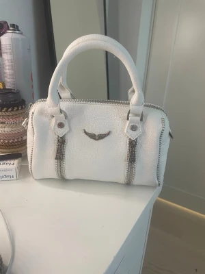 Vit handväska från Zadig & Voltaire - Snygg vit handväska från Zadig & Voltaire i strukturerat läder med silverfärgade detaljer och dubbla handtag. Väskan har dragkedjor på sidorna och en metallvinge framtill. Perfekt för dig som gillar en stilren och trendig look. Mått är 24x15cm