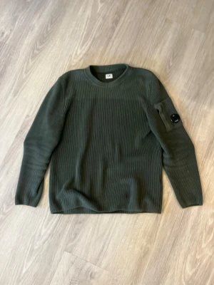 Mörkgrön stickad tröja från C.P. Company - Snygg mörkgrön stickad tröja från C.P. Company med rund halsringning och lång ärm. Tröjan har en unik detalj med en ficka och glaslins på ena ärmen. Perfekt för dig som gillar streetwear och stilrena plagg.