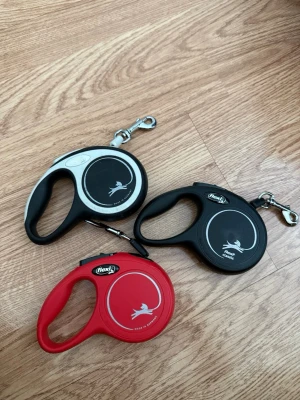 Tre Flexi hundkoppel i olika färger - Säljer tre Flexi hundkoppel i plast, ett svart, ett rött och ett svart/vitt. Alla har rundad form med handtag och metallhake för att fästa i halsband. Perfekt för promenader med hunden och smidiga att använda.