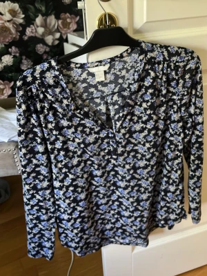 Blommig långärmad blus i blått - Snygg blus med småblommigt mönster i blått och vitt på svart botten. Blusen har v-ringning och långa ärmar. Tillverkad i ett mjukt och lätt tyg som känns skönt mot huden. Perfekt för dig som gillar en romantisk och trendig stil.