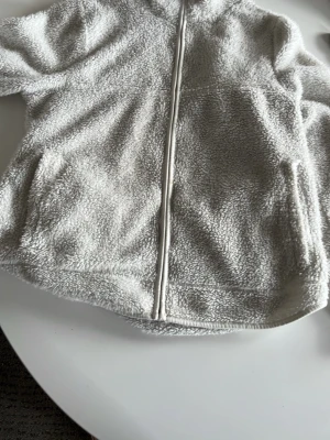 Ljusgrå teddyjacka med dragkedja - Mysig ljusgrå teddyjacka med hel dragkedja framtill och hög krage. Jackan har en fluffig och mjuk textur som ger en avslappnad look. Perfekt för kyliga dagar när du vill vara både varm och stilren.