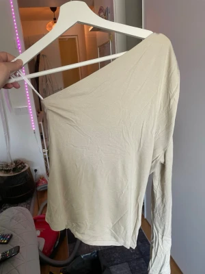 Beige oneshoulder topp - Trendig beige topp med oneshoulder-design och lång ärm. Perfekt för dig som vill ha en stilren och enkel look med en twist. Tunt och mjukt material som känns skönt mot huden.