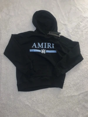 Svart hoodie Amiri Babyblue text - Svart hoodie från Amiri med stor huva och blått/vitt tryck framtill. Snyggt Amiri-logo och blå ränder över bröstet. Klassisk passform med ribbade muddar och känguruficka. Perfekt för en streetwear-look. Knappt använd och passar perfekt för S eller M de är 100% bomull så de sitter jätte skönt på huden, har fortfarande kvar själva shirttag som syns på bild 3. Hör av dig diskutera pris och kan postas direkt efter köpet.