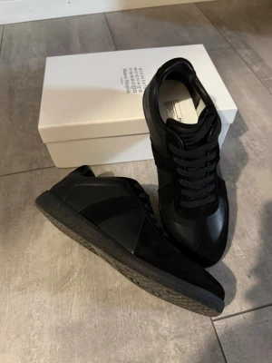  Maison Margiela  - Snygga svarta sneakers från Maison Margiela med klassisk låg siluett. Skorna har en mix av läder och mocka, svarta skosnören och en svart gummisula med unikt mönster undertill. Perfekta för dig som gillar stilrena och exklusiva sneakers.