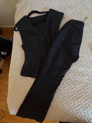 Svart set med väst och byxa - Stilren svart set med figursydd väst och matchande raka byxor. Västen har knappar framtill och en klassisk v-ringning. Perfekt för dig som vill ha en clean och tidlös look.