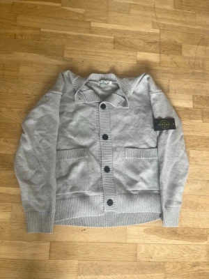 Grå kofta från Stone Island - 100% äkta kommer med online kvitto 100% bomull/ Snygg grå kofta från Stone Island med ribbstickade muddar, stora svarta knappar och två framfickor. Klassisk patch på ärmen och mjukt material som känns riktigt skönt. Perfekt för dig som gillar streetwear och vill ha något stilrent.