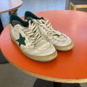 Golden goose ballstar - Säljer ett par klassiska vita ballstar sneakers med grön stjärndetalj på sidan och grön häl. Skorna har perforerad tåbox, vita skosnören och en gulnad sula för retrovibe. Tillverkade i läder och har låg profil.
