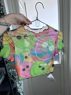 Färgglad mesh topp med smileys - Supercool mesh topp med korta ärmar och färgglatt smiley-mönster i grönt, gult, rosa och blått. Toppen har vågiga kanter vid hals, ärmslut och nederkant. Perfekt för dig som gillar statement-plagg och lekfull stil.