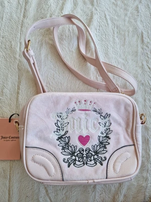 Ljusrosa axelväska Juicy Couture - Söt ljusrosa axelväska från Juicy Couture i sammet med broderad logga, hjärta och krona framtill. Väskan har justerbar axelrem och guldfärgade detaljer. Perfekt för dig som gillar en trendig och feminin stil.