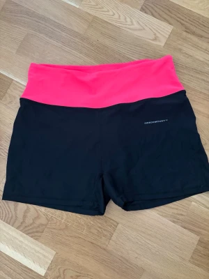 Svarta träningsshorts Swedemount - Svarta träningsshorts från Swedemount med bred, neonrosa midja. Shortsen är stretchiga och har en hög midja som sitter skönt vid aktivitet. Perfekta för gymmet eller löprundan.