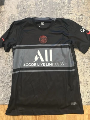 Svart PSG matchtröja från Nike - Snygg svart PSG matchtröja från Nike med röd Paris Saint-Germain-logga på bröstet och vit sponsorlogga 'Accor Live Limitless' framtill. Tröjan har korta ärmar, rund hals och är gjord i ett lätt, sportigt material perfekt för träning eller match.