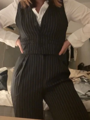 Svart randig väst med knappar - Snygg svart väst med vita tunna ränder och tre knappar framtill. Klassisk v-ringad modell med fickdetaljer.