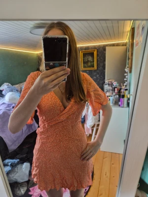 Orange omlottklänning från Kookai - Superfin orange kort omlottklänning från Kookai med vita prickar. Klänningen har volangkant nertill, v-ringning och korta ärmar. Perfekt för sommarens alla tillfällen och ger en fräsch och trendig look.