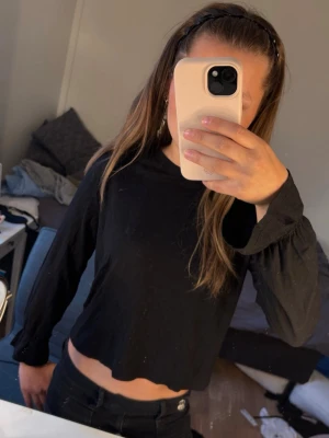 Svart långärmad crop top - En svart långärmad crop top med rund hals och vågig kant nertill. Toppen har en enkel och stilren design, perfekt att matcha med höga jeans eller kjol. Materialet ser ut att vara mjukt och stretchigt, troligen bomull eller bomullsblandning.