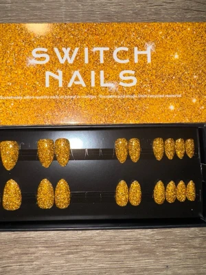 Switch Nails Gold Flash press-on naglar - Glittriga press-on naglar i modellen Gold Flash från Switch Nails. Naglarna har en kort mandelform och är täckta av guldigt glitter. Tillverkade av återvunnet material för en hållbar look. Perfekt för dig som vill sticka ut med snygga, gnistrande naglar. Säljes utan tillbehör.