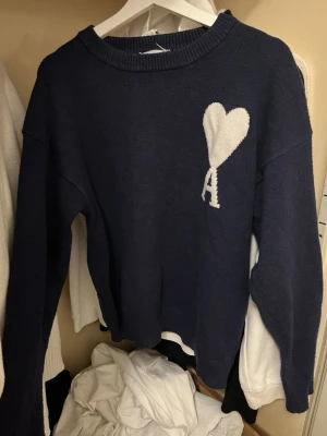 Mörkblå stickad tröja Ami Paris - Mörkblå stickad tröja från Ami Paris med vit hjärtlogga och bokstaven A på bröstet. Tröjan har rund halsringning, långa ärmar och ribbade muddar. Perfekt för dig som gillar stilrena och trendiga plagg.