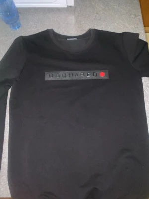 Svart långärmad topp från Dsquared2 - Snygg svart långärmad topp från Dsquared2 med ett diskret tryck på bröstet och en liten röd detalj. Klassisk rund halsringning och rak passform. Perfekt för dig som gillar stilrena plagg med en twist.