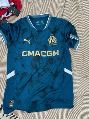Marseille matchtröja blå Puma - Snygg blå matchtröja från Puma för Olympique de Marseille med guldiga detaljer och logga. Tröjan har korta ärmar, vit krage och vita detaljer på ärmarna. 