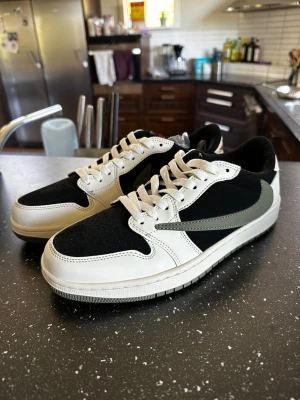 Nike Air Travis scott Jordan 1 Low svart/vit - Snygga Travis scott Nike Air Jordan 1 Low sneakers i svart och vitt med grå swoosh. Ovandel i mocka och läder, klassisk AJ1-design och röda detaljer på hälen. Skorna kommer med extra skosnören i olika färger. Perfekta för dig som gillar streetwear och vill sticka ut.