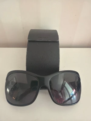 Svarta Prada solglasögon med etui - Snygga svarta solglasögon från Prada med breda bågar och mörka glas. Modellen har en klassisk, lite större form som ger en cool look. Kommer med originaletui i svart. Perfekta för att lyfta din stil under soliga dagar.