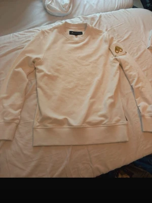 Vit sweatshirt från Moose Knuckles - Snygg vit sweatshirt från Moose Knuckles med klassisk passform och ribbade muddar. Tröjan har en guldfärgad logga på ärmen och är tillverkad i mjukt bomullsmaterial. Perfekt för en clean och stilren look.
