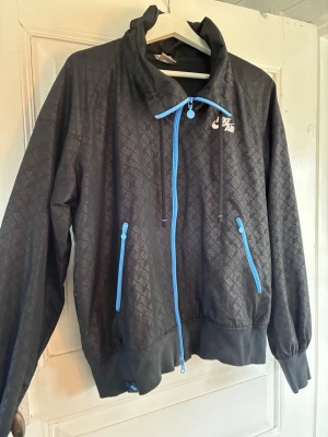 Svart Nike Air vindjacka XL - Nike Air vindjacka i svart med diskret mönster och blå dragkedjor. Jackan har hög krage, snörning i huvan och två fickor med dragkedja. Tillverkad i syntetmaterial och har en sportig look. Passar snarare L.