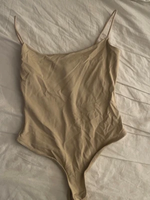 Beige body med smala axelband - Snygg beige body med smala axelband och rundad halsringning. Tillverkad i ett mjukt och stretchigt material, perfekt för lager-på-lager. Praktisk knäppning i grenen för enkel användning.
