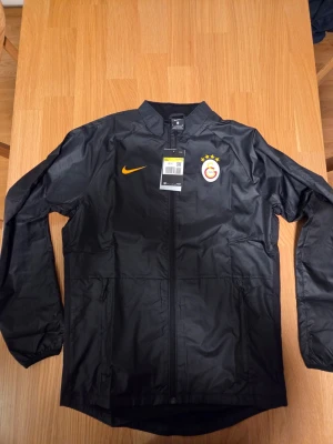 Nike Galatasaray academi repel dry-fit träningsjacka, storlek S - Svart Nike-jacka med Galatasaray-logotyp på bröstet och en stor orange Nike-swoosh på ryggen. Jackan har hel dragkedja framtill och sidofickor, varav en med dragkedja. Den har även perforerade detaljer längs sidorna.  Artikelnummer: DA9791-010