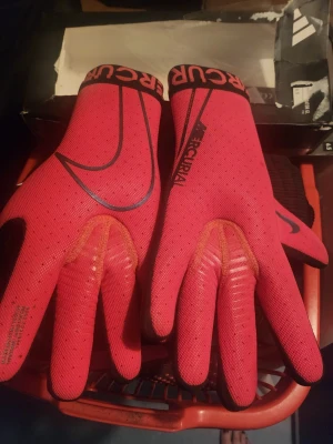 Nike Mercurial målvaktshandskar röd/svart - Snygga Nike Mercurial målvaktshandskar i rött och svart med svart Nike-logga. Ovansidan är röd med svarta detaljer och insidan har greppvänligt svart material. Perfekta för fotbollsmatcher och träning. Sitter tajt och ger bra känsla i spelet.