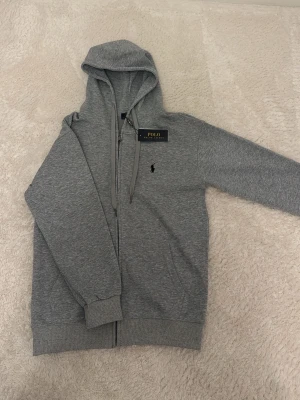 Grå hoodie från Polo Ralph Lauren - Snygg grå hoodie med dragkedja från Polo Ralph Lauren. Klassisk design med huva och dragsko, ribbade muddar och midja samt den ikoniska lilla broderade loggan på bröstet. Perfekt för en chill och stilren look.