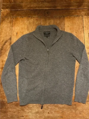 Grå merino/kashmir ziptröja från Savile Row Company - Grå ziptröja i merinoull och kashmir från Savile Row Company. Tröjan har hel dragkedja, ståkrage och ribbade muddar. Perfekt för lager-på-lager och ger en clean look till din outfit.