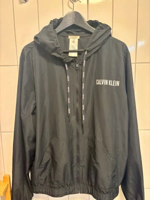 Calvin Klein vindjacka - Snygg svart vindjacka från Calvin Klein med dragkedja framtill, huva med justerbara snören och två fickor. Jackan har vit logga på bröstet och snören med Calvin Klein-tryck. Tillverkad i lätt och smidigt material, perfekt för blåsiga dagar.  Inköpspris 1299